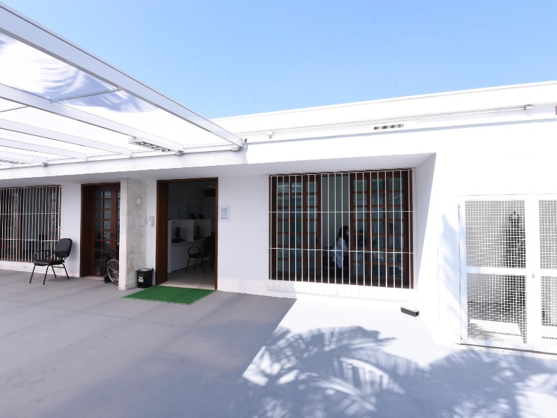 A fachada da casa é branca com duas janelas e duas portas grandes. Ao lado de uma das portas há uma cadeira preta e pela janela é possível ver uma mulher de jaleco branco